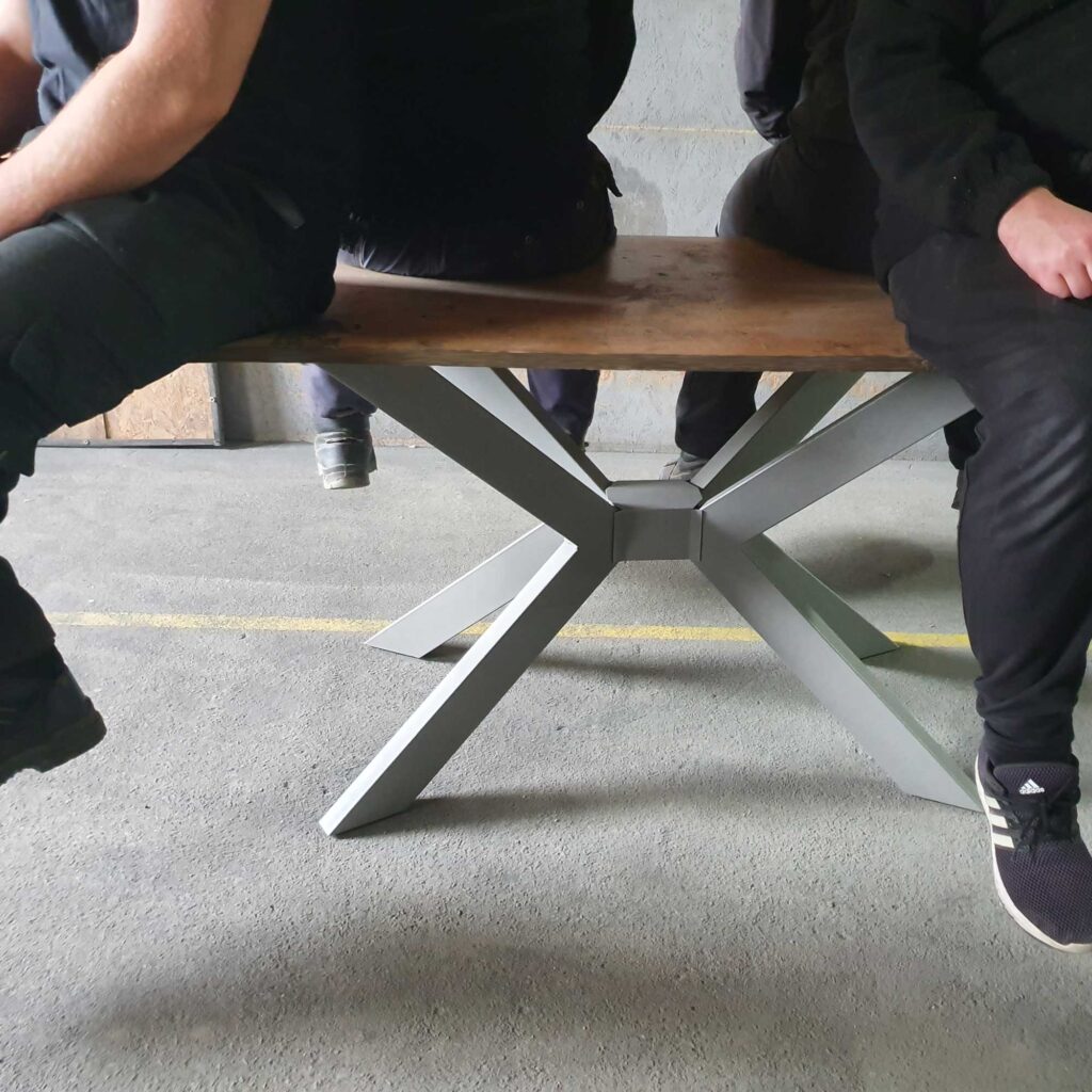 Vier Personen sitzen auf dem 4V Tischgestell und testen die extreme Tragfähigkeit für schwere Tischplatten.