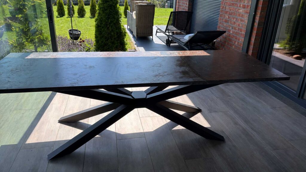 Modern black rectangular table with crisscross metal legs on a wooden patio, overlooking a garden.