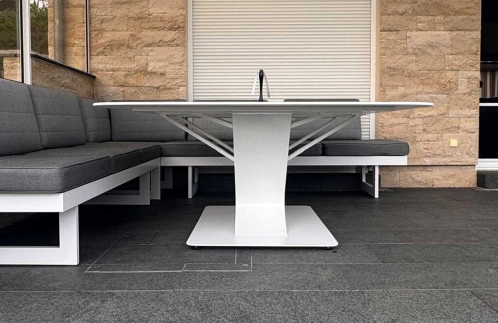 Outdoor patio scene with a white modern table and gray cushioned L-shaped sofa against a stone wall.