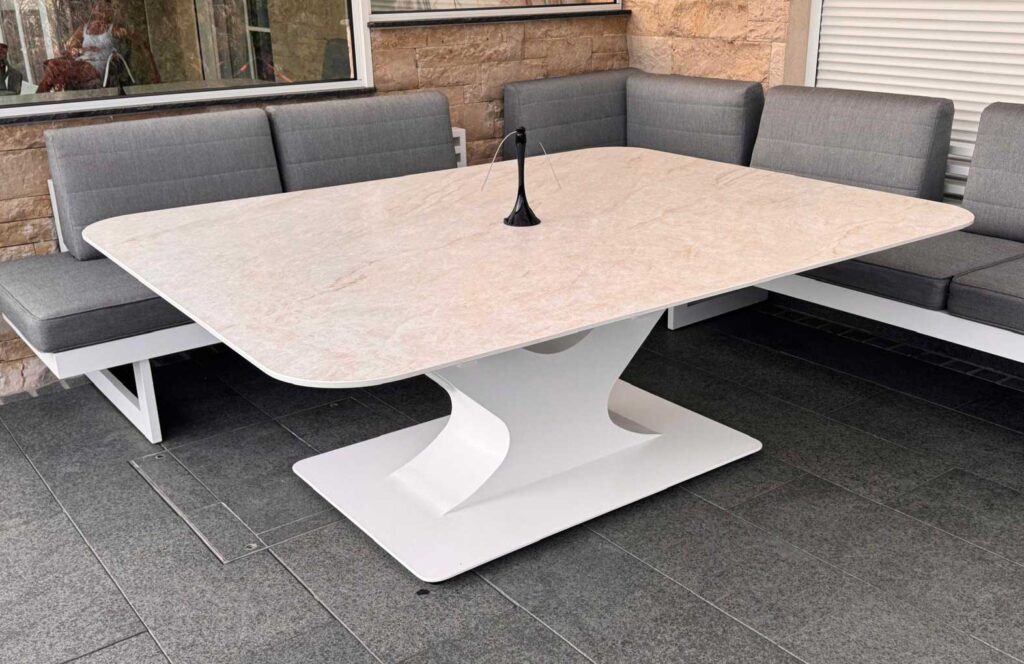 Outdoor patio seating area with a large white pedestal table and gray cushioned benches along a stone wall.