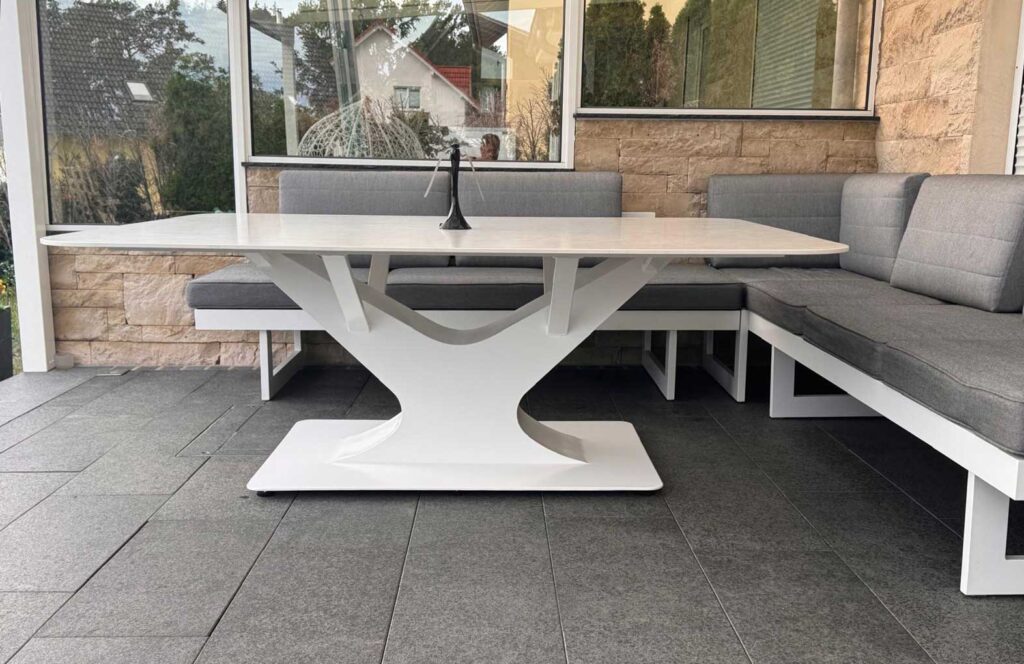 Outdoor patio dining area with a white sculptural pedestal table and gray cushioned corner bench along a stone wall.