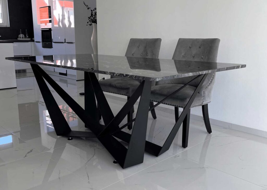 Modern dining table with a glass top and black geometric legs, paired with gray upholstered chairs in a bright kitchen.